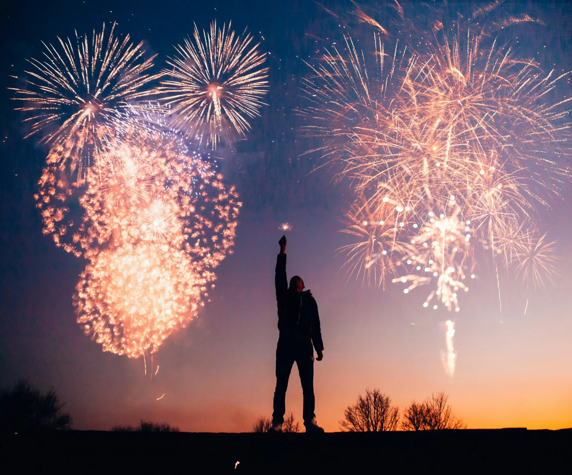 Man holding a sparkler in his hand with fireworks going off in the background.